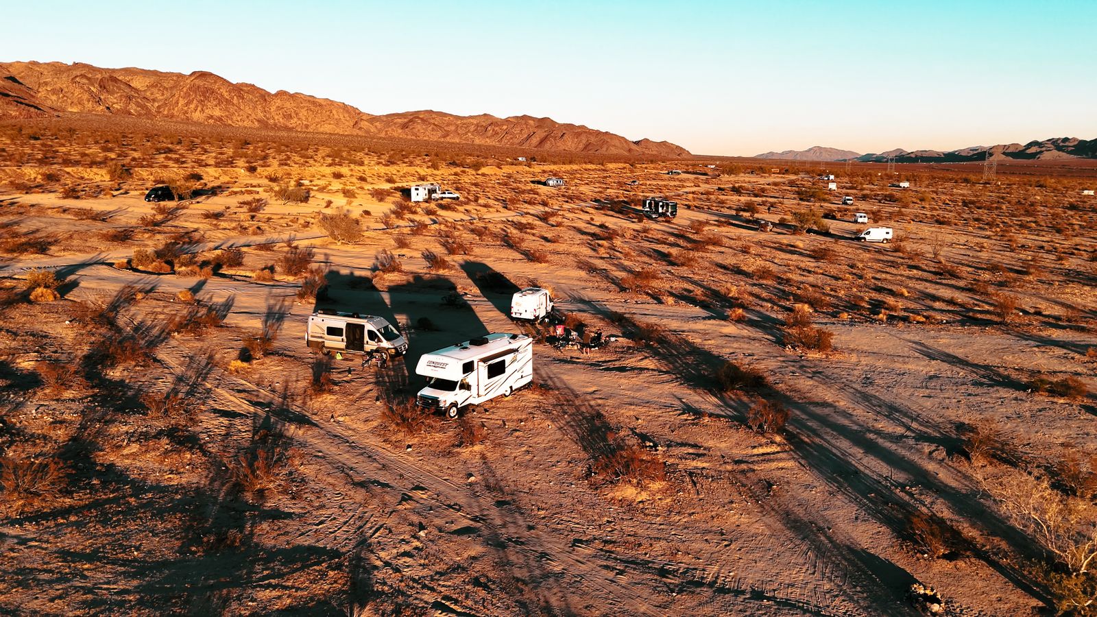 Motorhome and travel trailer camped in open desert as the sun drops behind distant mountains